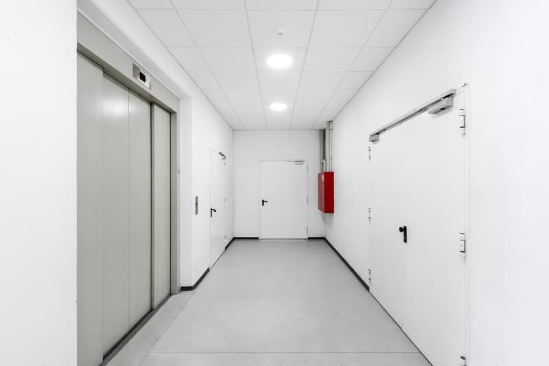 wide hallway, white walls, white floor, elevator shaft, door at the end, fire extinguisher
