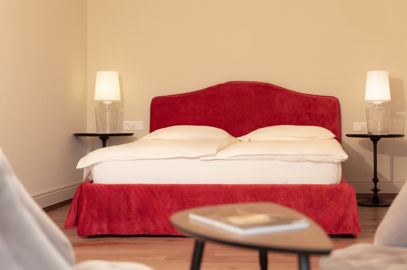 Double bed with red headboard and bed skirt, white pillows, nightstands with lamps and power outlets, wooden coffee table with books, wooden floor, white walls