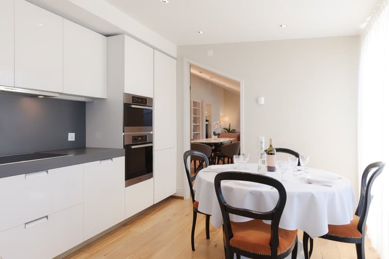 White kitchen, wooden floor, large dining table, wine glasses, bottles, open doorway to dining area.