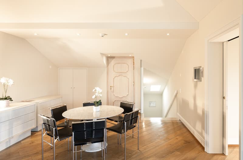 Dining area with a wooden table and modern chairs. White walls and cabinets, flower vases on a table and shelf, and a glass door leading to a staircase.
