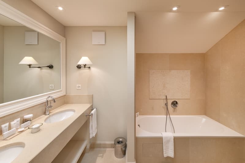 modern bathroom, beige tiles, double sinks, a bathtub, white towels, wall-mounted lights, countertop with toiletries