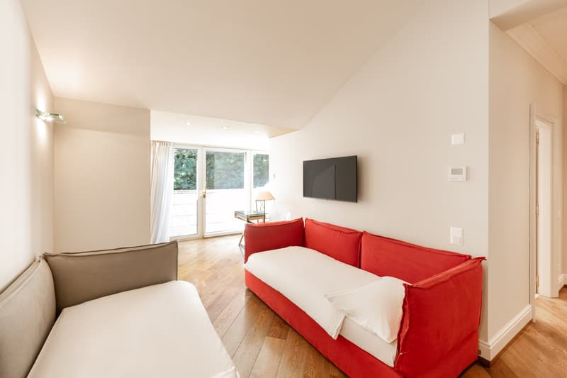 Two comfortable couches, hardwood floor, windows with white curtains, television, side table with lamp, white walls, wall lights