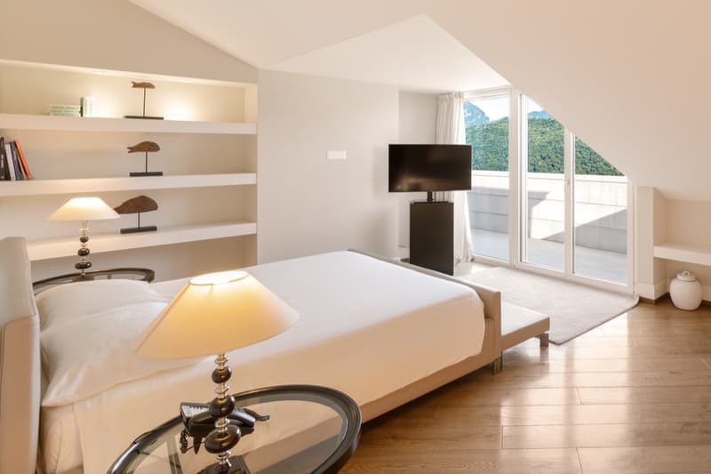 A bedroom with a bed, two lamps, a rug, and a television. Features include large windows, a balcony, and a view of the mountains.