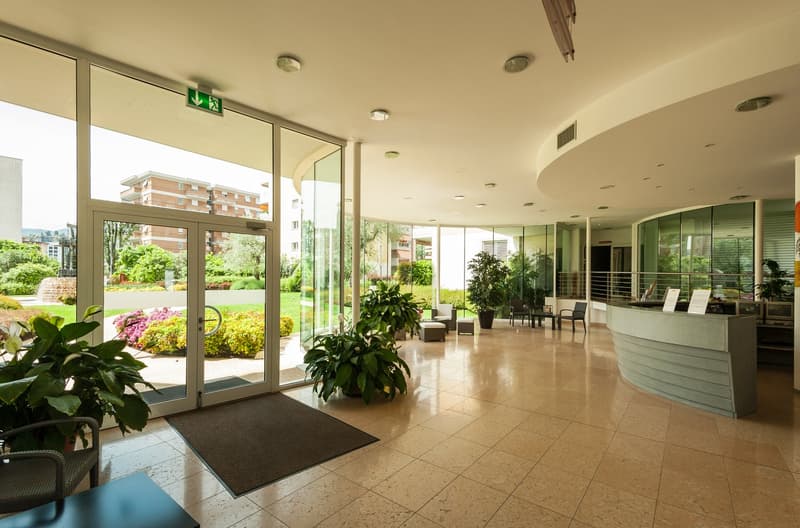 Spacious lobby with large windows overlooking a garden, tiled flooring, potted plants, and a reception desk