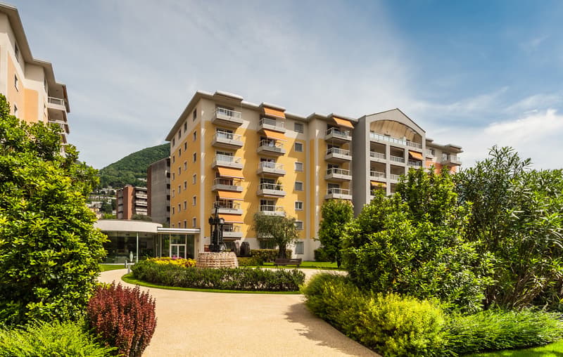 Multi-story apartment building with yellow and gray exterior, balconies, surrounded by lush greenery and landscaping, with a fountain or water feature in the foreground