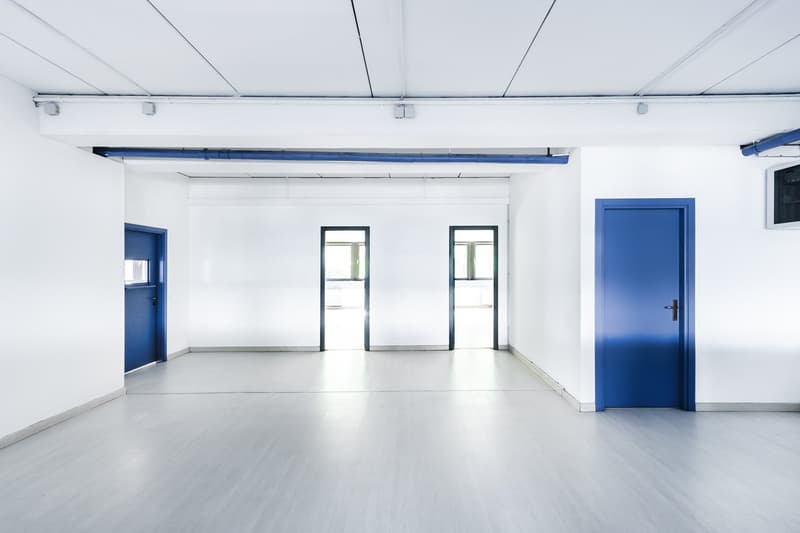 Large open room with white walls, concrete floor, and blue accents. Two doorways and windows are visible. The room appears to be a commercial or office space with minimal furnishings.