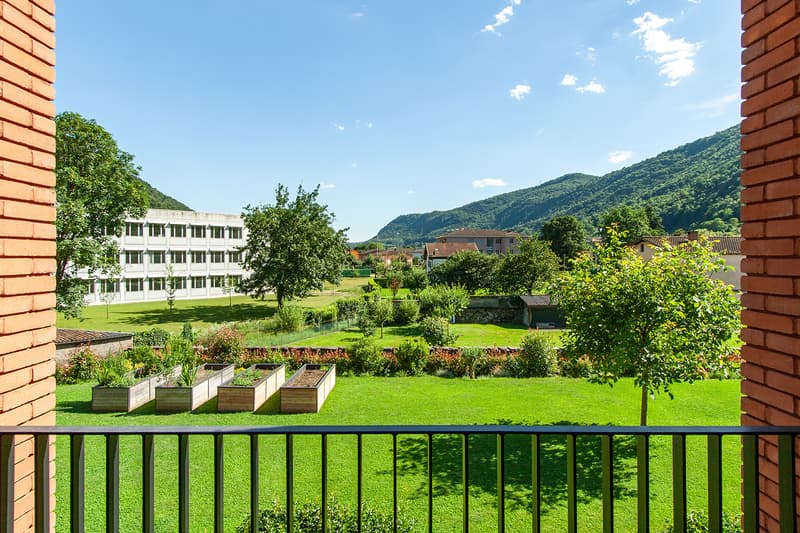The image shows a residential building surrounded by a lush, green landscape. The building has multiple floors and is made of red brick. There is a balcony or terrace visible, providing a view of the scenic outdoor area. The grounds include a grassy lawn,