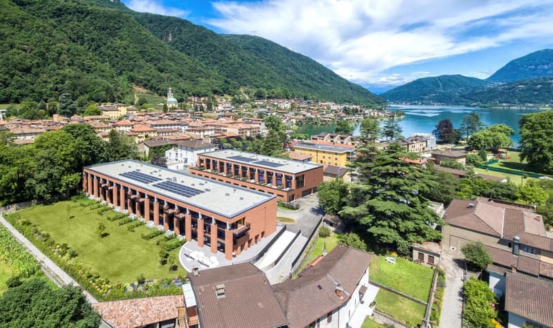 Large modern brick building with solar panels on the roof, surrounded by a lush green landscape and overlooking a lake and mountains in the background. The building appears to be situated in a picturesque town or village nestled in the mountains.
