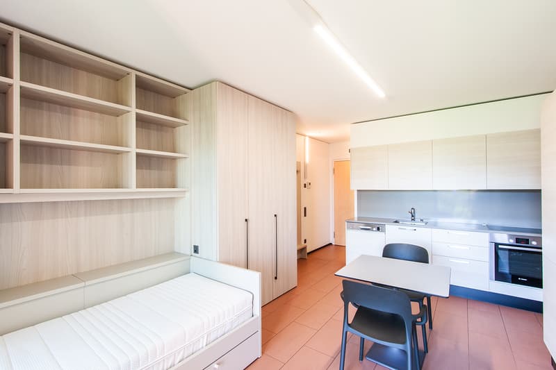 The image shows a studio apartment with a kitchenette, dining area, and a built-in storage unit. The apartment has a modern and minimalist design, with white walls, wooden shelves, and tile flooring. The kitchen area features a dishwasher and other applia