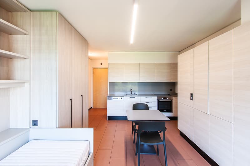 Open-plan studio apartment with kitchen, dining area, and sleeping area. The kitchen features built-in appliances including a dishwasher. The living space has built-in shelving and storage along the walls. The flooring is tiled throughout.