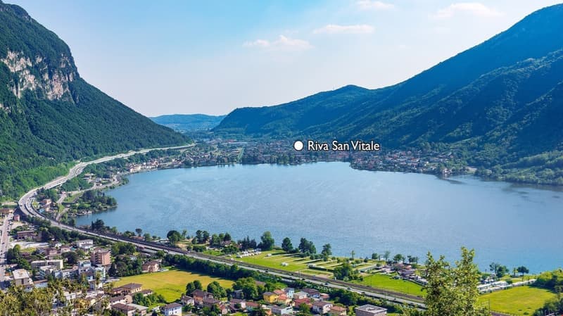Aerial view of Riva San Vitale, a town located on the shores of a large lake surrounded by mountains. The town is situated along the lakefront with residential and commercial buildings, and there is a road running along the lake. The landscape is scenic w