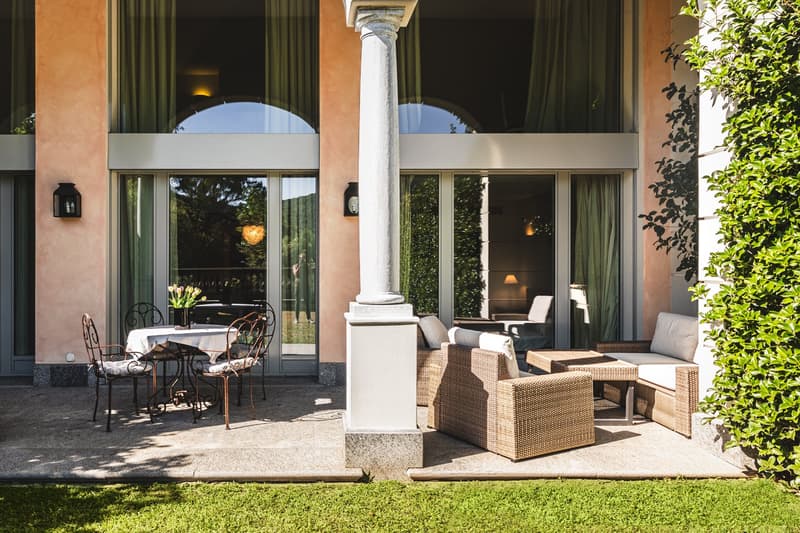 Outdoor patio with wooden chairs, table, wicker sofa, green lawn, glass doors, stone column, plants, wall-mounted lamps