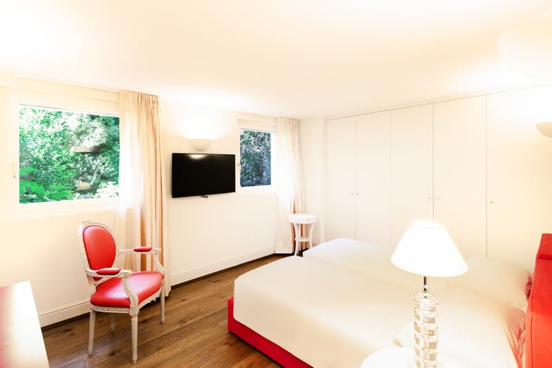Minimalist style bedroom with double bed, television, chair, lamp, white wardrobe, nightstands, and two large windows with curtains