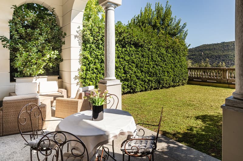 Terrace with dining table, wicker chairs, flowers in a vase, green lawn, hedge, mountain in the distance.