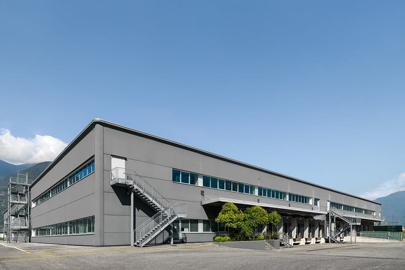Large industrial or commercial building with multiple floors, gray exterior, multiple entrances and staircases, and a parking area in front of the building.