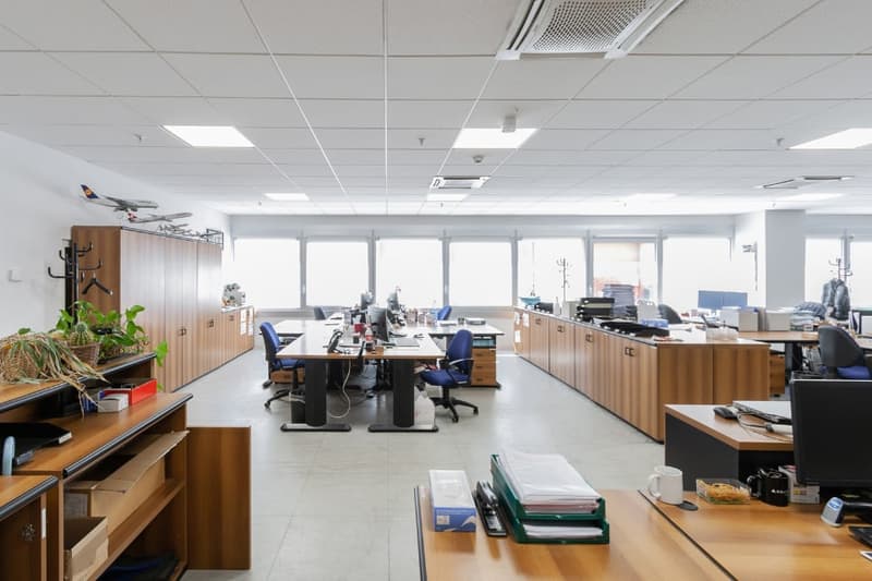 Open office space with cubicles, desks, computers, and office equipment. The room has large windows, a tiled floor, and a suspended ceiling with lighting fixtures.