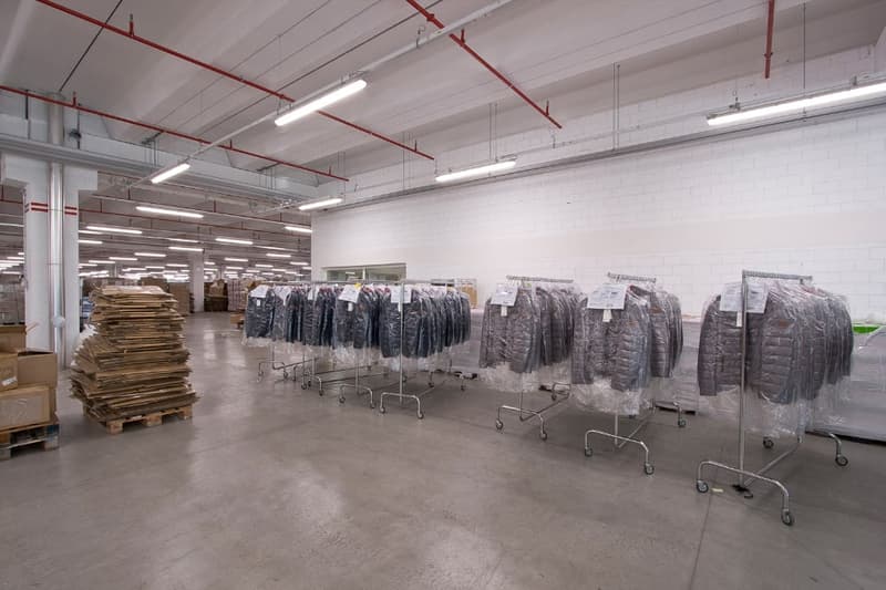 Large industrial warehouse space with rows of clothing racks, stacks of cardboard boxes, and overhead lighting. The space appears to be used for storage and distribution of clothing items.