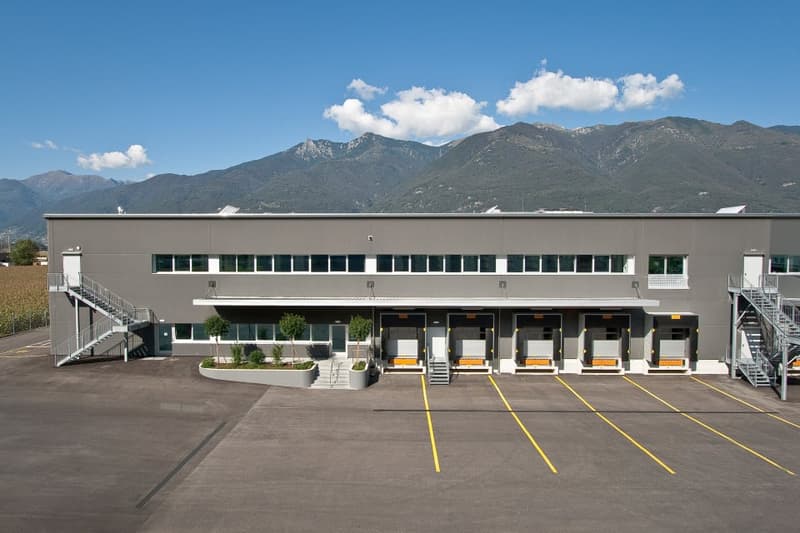 A large commercial building with multiple loading docks and parking spaces. The building has a gray exterior and is surrounded by mountains in the background.
