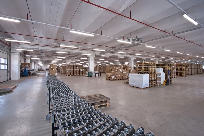 Large industrial warehouse space with high ceilings, rows of shelving and pallets stacked with goods, and a fleet of shopping carts or trolleys in the foreground.