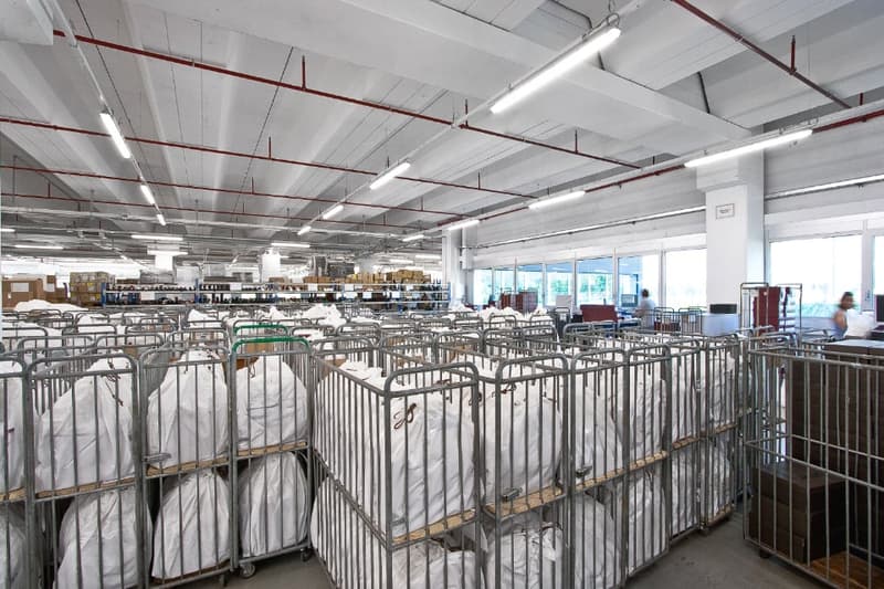 Large industrial warehouse space with rows of shelving units and storage containers, high ceilings with exposed ductwork and lighting fixtures