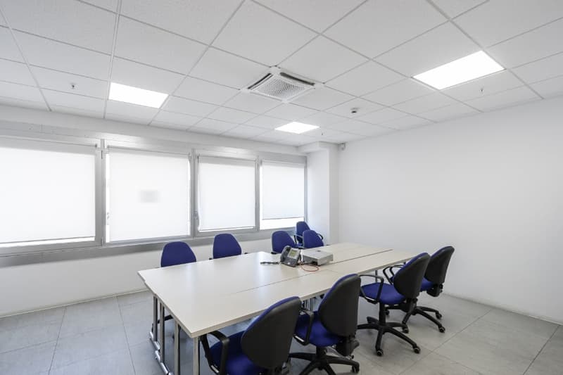 The image shows a modern, well-lit office space with a large conference table surrounded by blue office chairs. The room has a clean, minimalist design with white walls, a tiled floor, and recessed lighting fixtures in the ceiling. The large windows provi