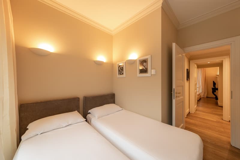 Bedroom with two twin beds, white linens, beige walls, wall lights, two picture frames, and an open door leading to another room.