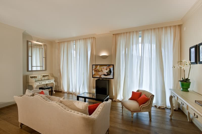 beige sofa, beige chair, TV on cabinet, small glass table, large white curtains, mirror, white writing desk