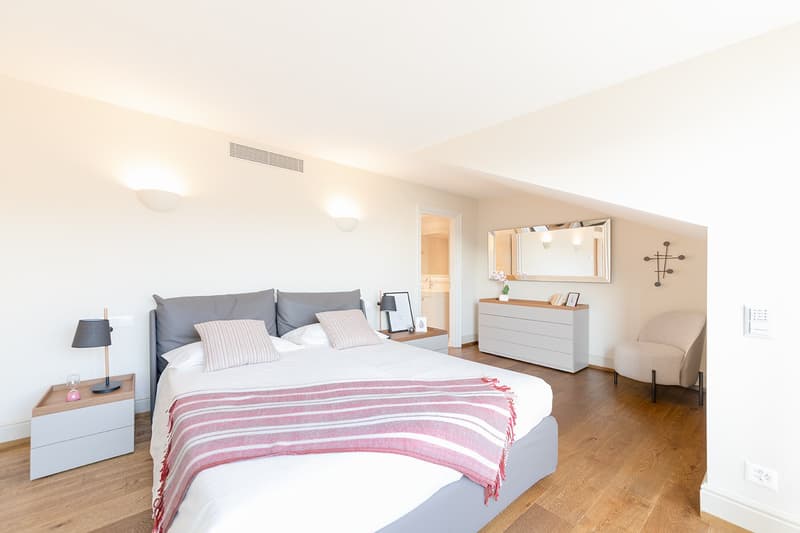 Master bedroom with a queen-size bed, wooden floors, white walls, nightstands, lamp, mirror, dresser, armchair, and decorative items.