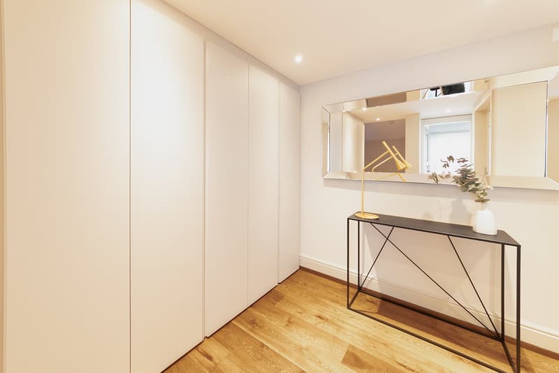 Modern room with white walls and wooden floor, a mirror with a lamp and plant on the wall, a black console table, and a set of white built-in closets.