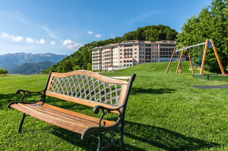 Bench on the lawn, behind the bench there is a large hotel with many floors, in the background a green mountain and a blue sky
