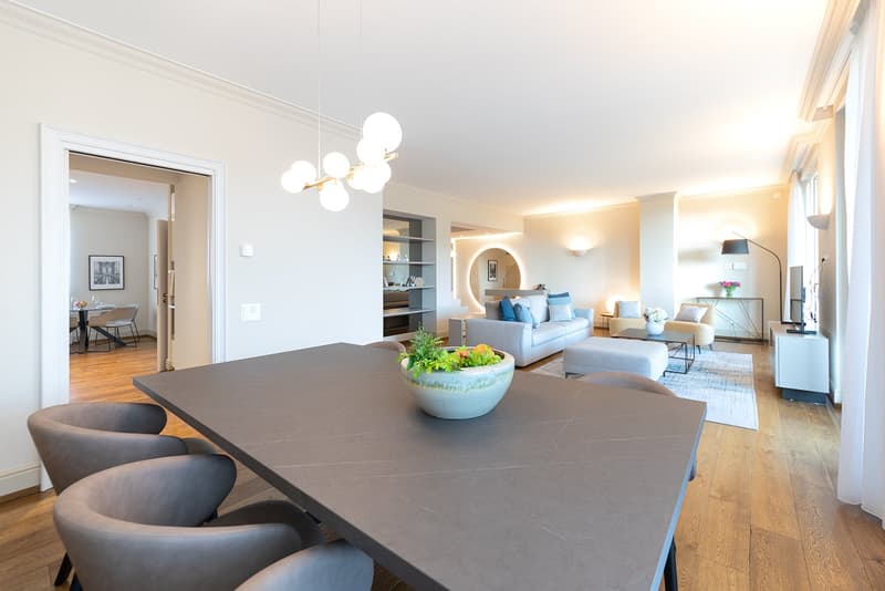 Large dining table, wooden floors, chandelier, round plant pot, gray chairs, living room with couch and ottoman