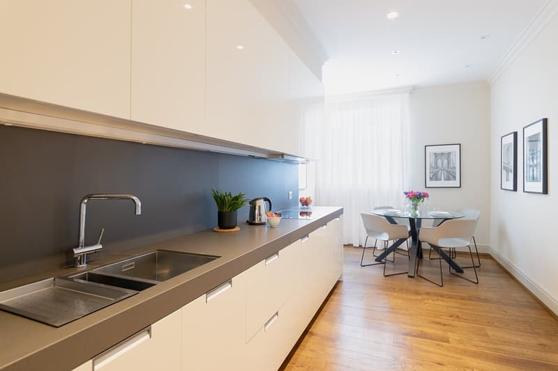 Modern kitchen with white cabinets, gray countertop, sink with stainless steel faucet, black kettle, plant, stainless steel dishwasher, round dining table, chairs, white curtains