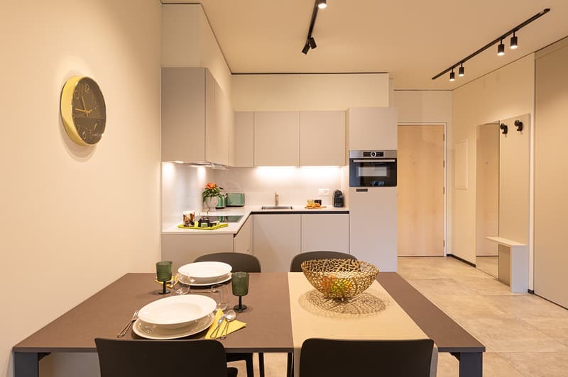 Modern dining room with wooden table, chairs, ceramic tableware, dining basket, wine glasses, utensils, contemporary decor, kitchen with microwave, coffee machine, and dining space.
