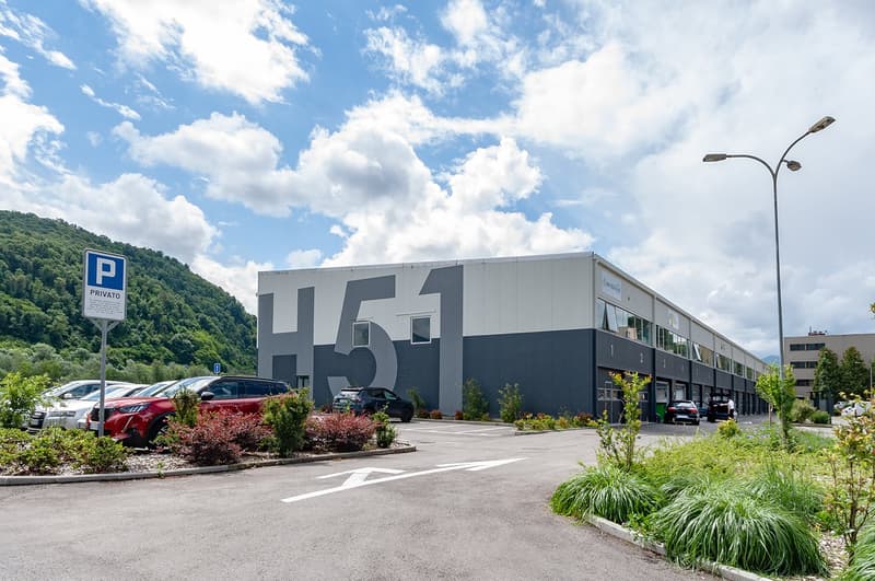 Large building with a parking lot in front, number 151 on the side, surrounded by greenery and a mountain in the background.