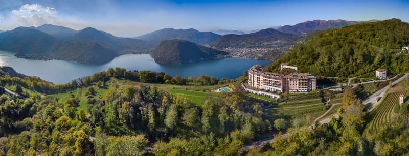 Large building with a green roof, surrounded by trees, mountains, and a lake