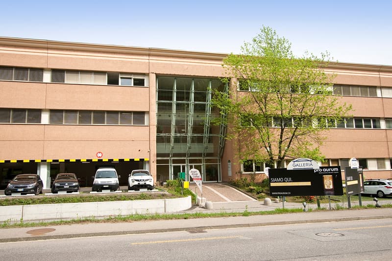 A pink building with glass windows and doors, several cars parked in front, a tree in front, and a signboard on the right side.