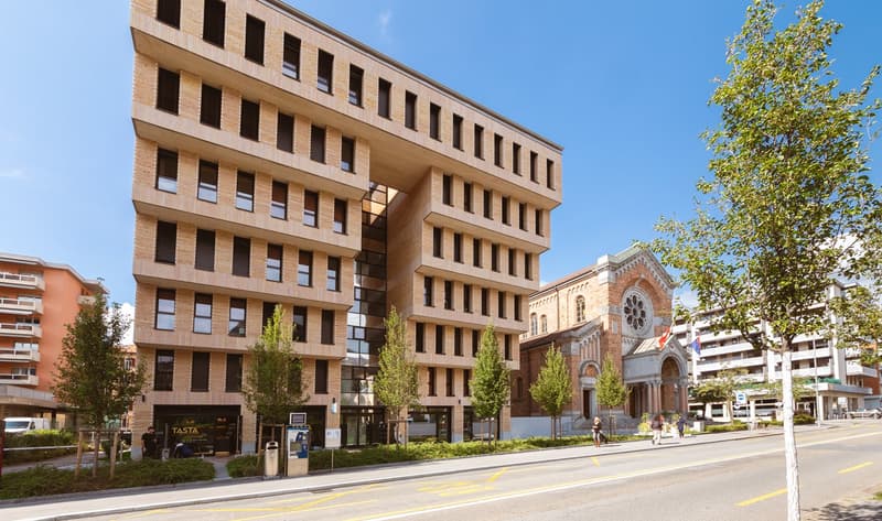 Modern building with brick facade, multiple windows, and a church in the background.