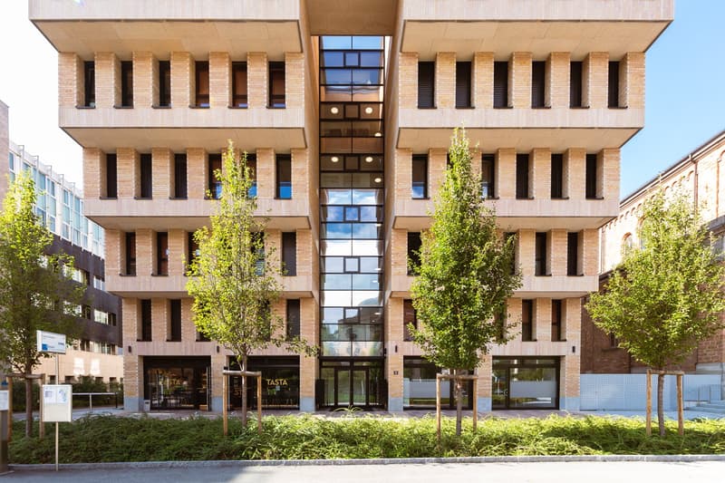 Modern multi-story building with brick facade, large glass windows, central atrium, and entrance with glass doors.