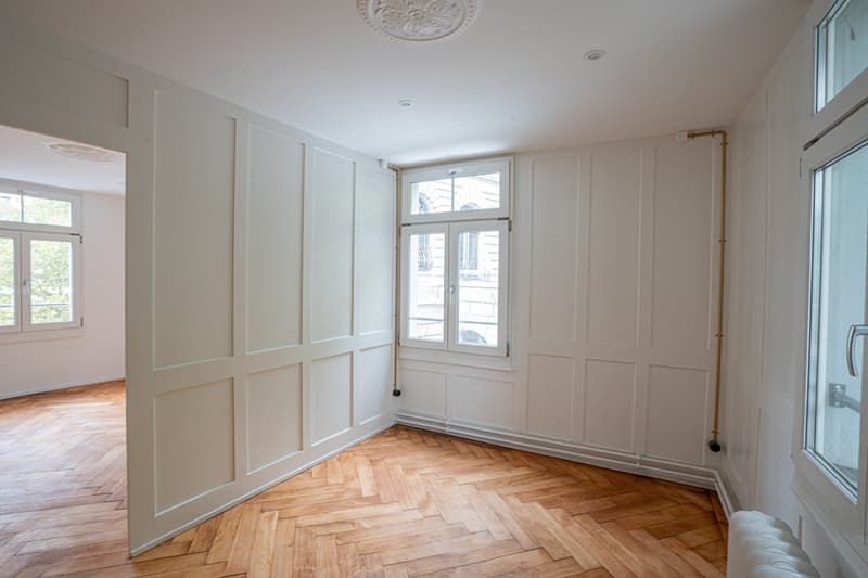 Empty room with wooden parquet floors, white walls, and ceiling with a decorative medallion, two windows with doors, one leading to another room, and an old-style radiator