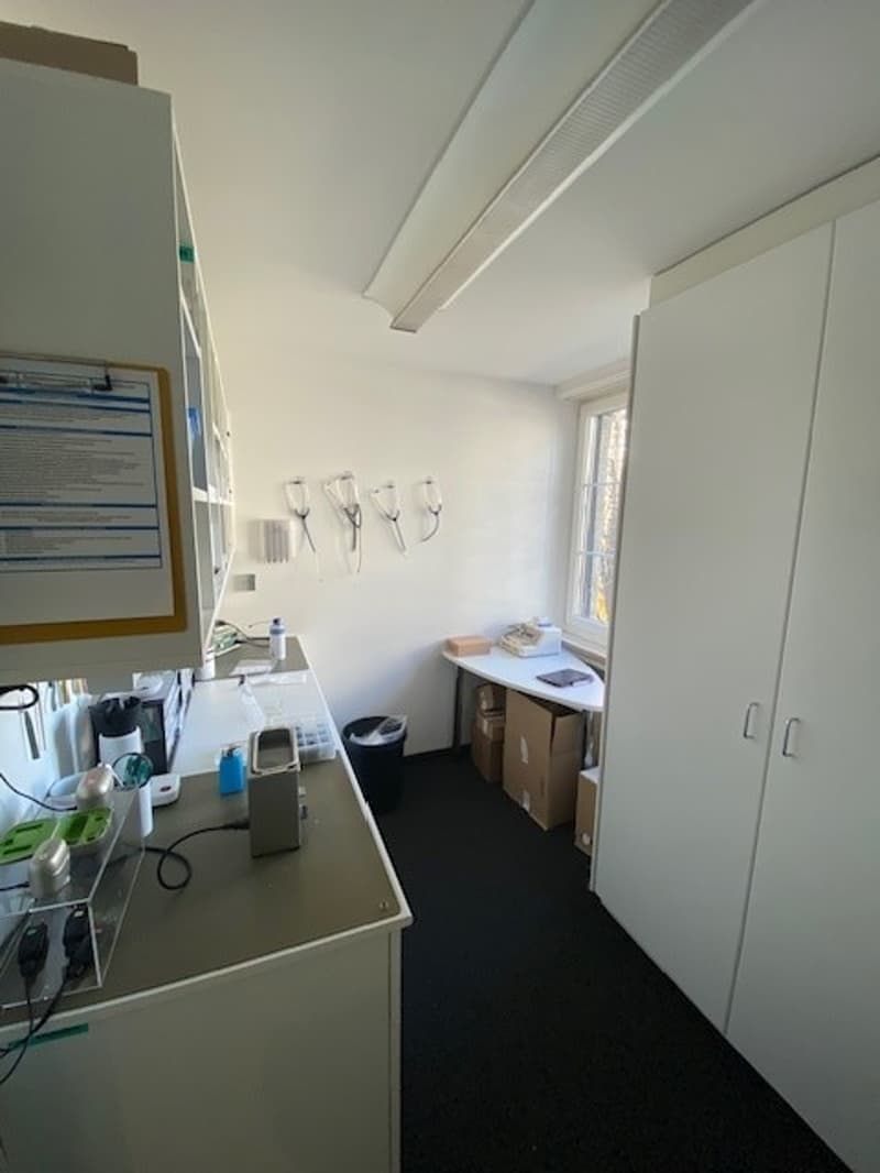 White-walled room with a desk, boxes, and various medical equipment. The ceiling has an air vent, and there's a trash bin, a window with blinds, and wall-mounted shelves. The floor is black and covered with carpet. A small refrigerator and other electroni