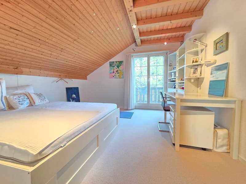 ample bedroom with wooden ceiling, bed with drawers, white bedding, desk, chair, shelf, rug, balcony door, paintings on the wall