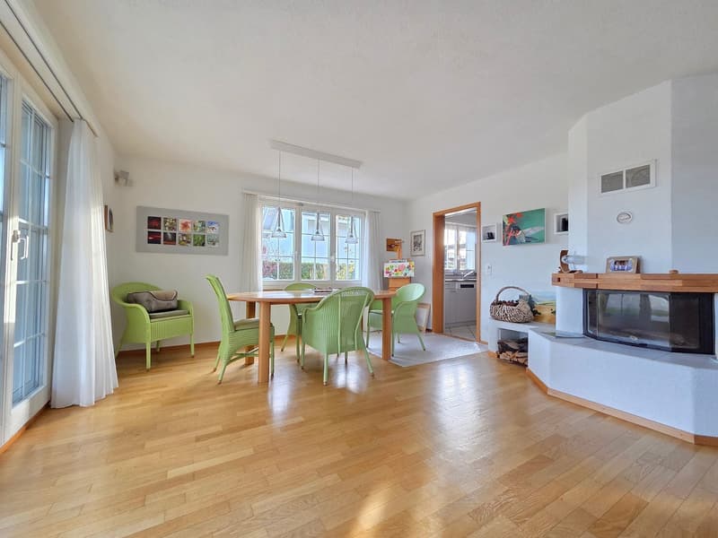 Dining area with wooden table, green chairs, white curtains, fireplace, large windows, pendant lamps