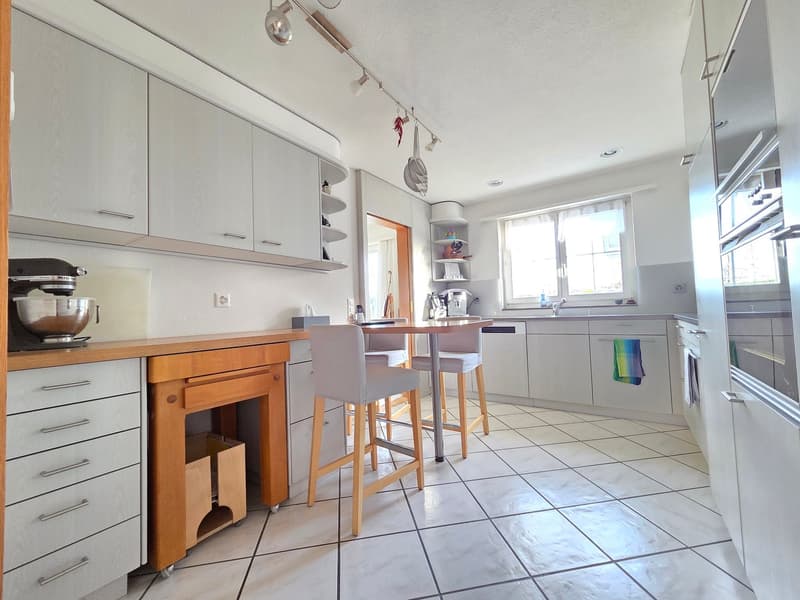 kitchen with large counter, wooden barstools, white cupboards, white tiled floor, island table, microwave, sink, dishwasher, cabinet for storage, refrigerator, electric kettle, kitchenware hanging from lamp holder