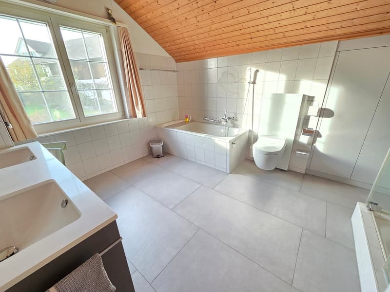 A white bathroom with ceramic tiles, two sinks, a bathtub, a toilet, a shower, and a trash can. The room also features a window with curtains and a wooden ceiling.