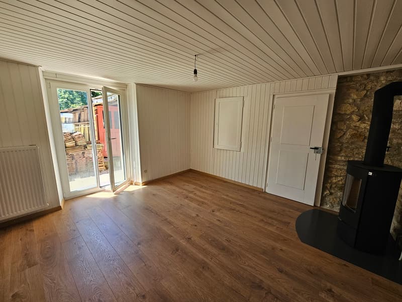 This image shows a spacious room with a wooden floor, white paneled walls, and a sliding glass door leading to an outdoor area. The room also features a wood-burning stove or fireplace. The overall aesthetic is rustic and cozy.
