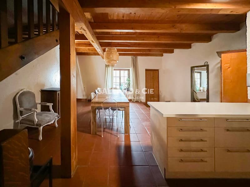 large wooden beams, terracotta tiles, dining area with wooden table and chairs, kitchen area with drawers, staircase, mirror, lamp, windows, and a window seat