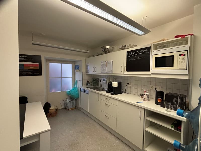 A well-equipped kitchen with white cabinets, a countertop, a sink, and an integrated coffee machine. It has an oven, a microwave, and other kitchen appliances on the shelves. The room also has a window, a table, and some storage boxes.