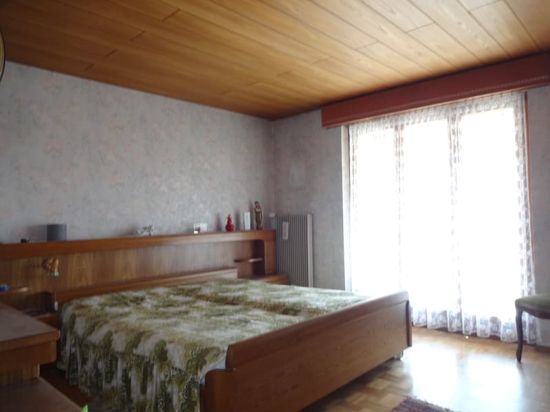 wooden ceiling, bed with wooden headboard, floral bedspread, two nightstands, large window with white curtain
