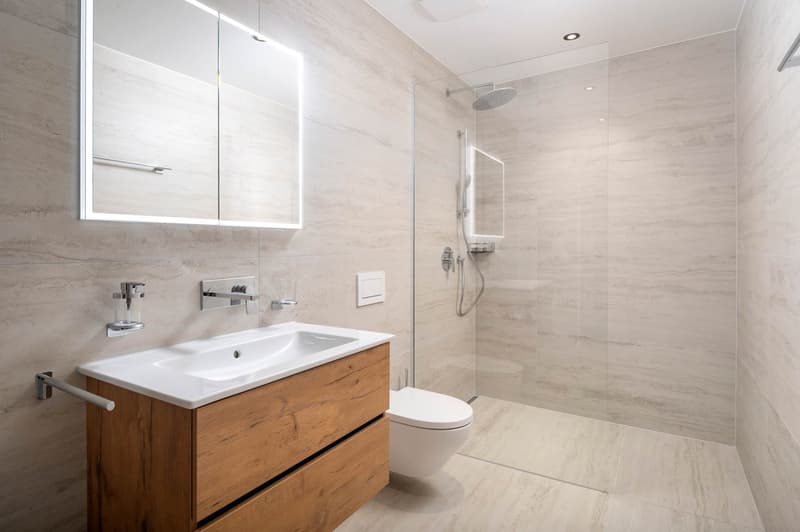 modern bathroom with white sink, wooden cabinet, glass shower enclosure, white toilet, mirror with lighting