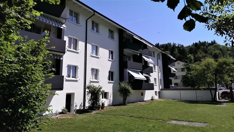 Three-story white building with multiple balconies, trees, and a parking lot in front